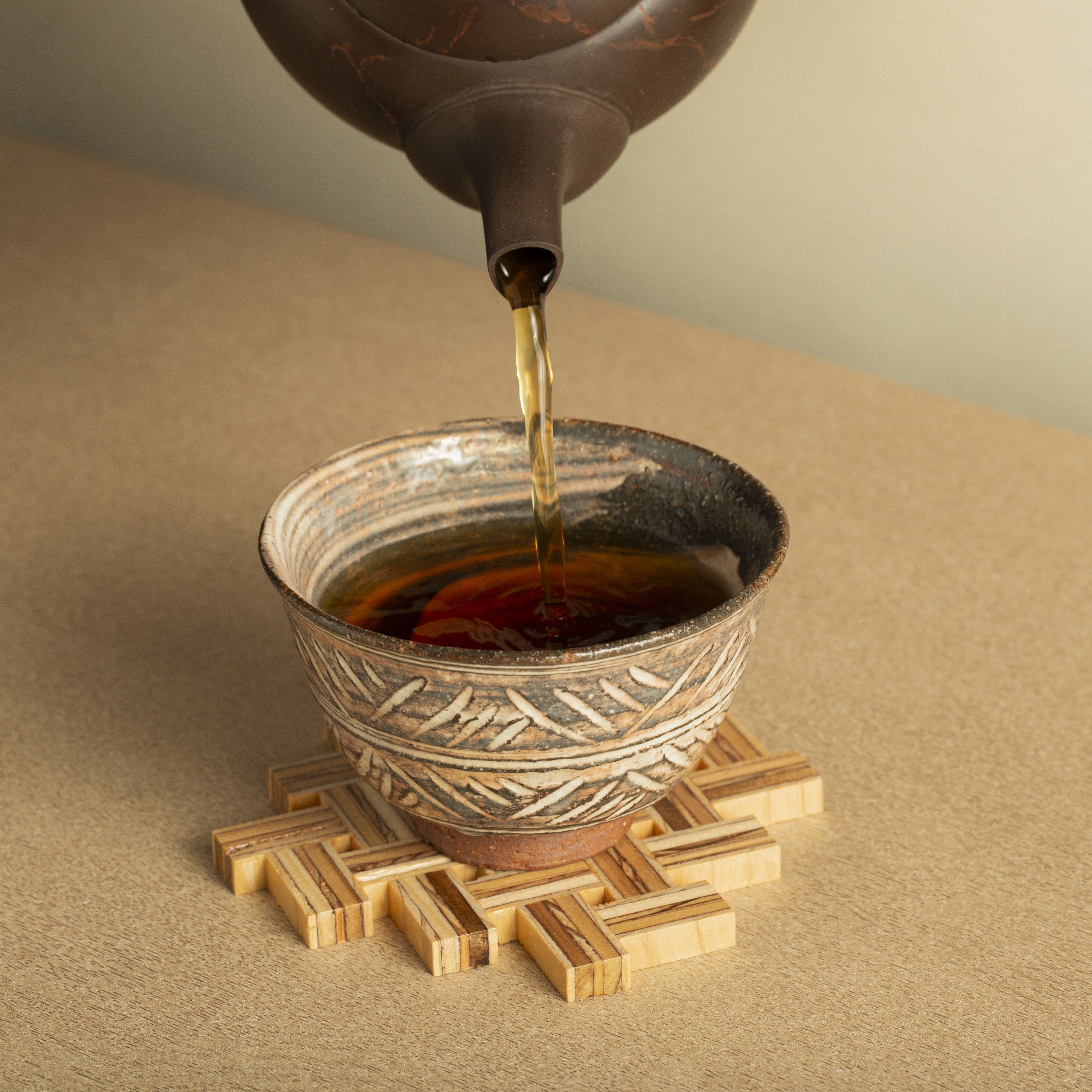 Tea being poured from a teapot into a ceramic cup on a textured surface.