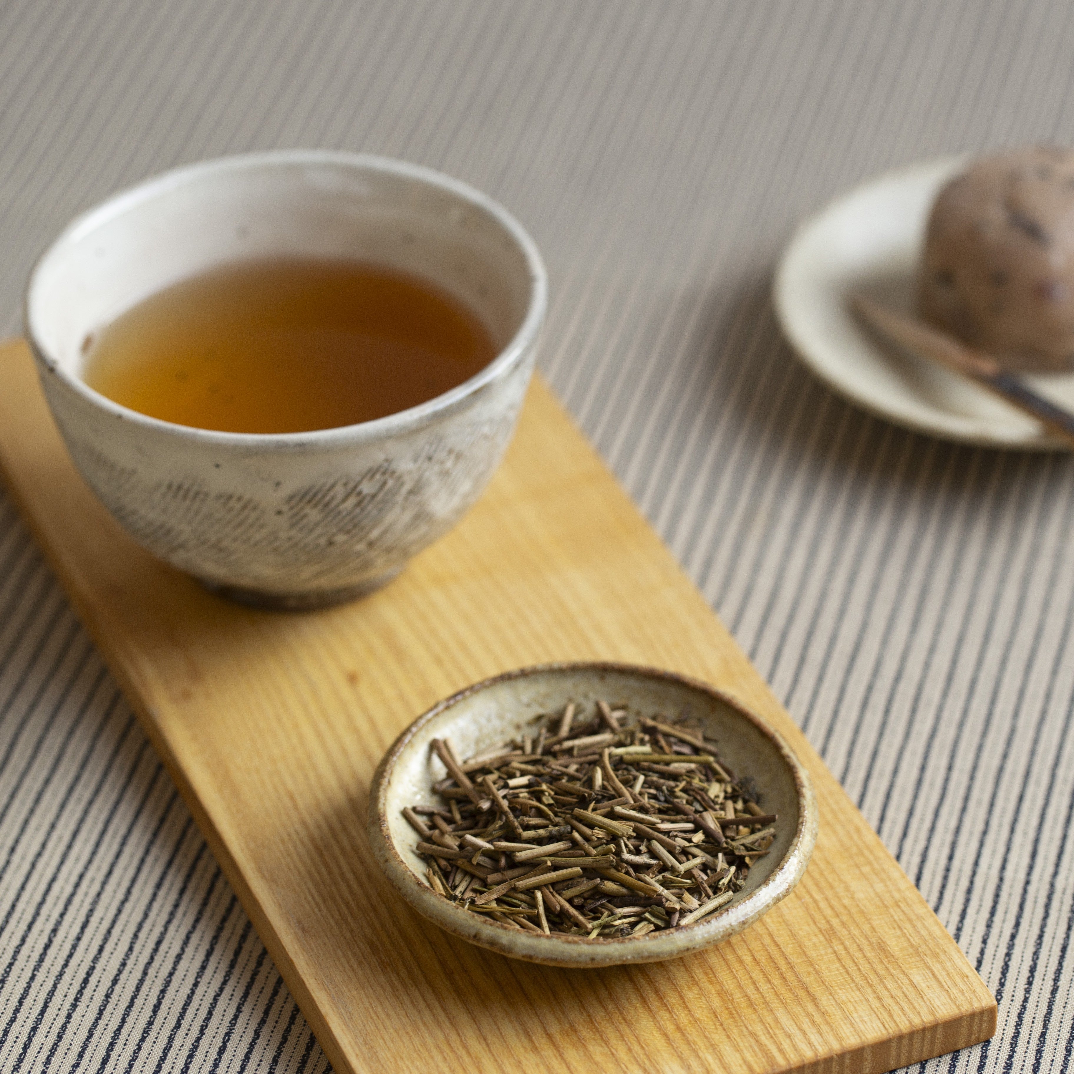 Tea cup with brewed tea and a bowl of hojicha loose tea leaves on a wooden tray.