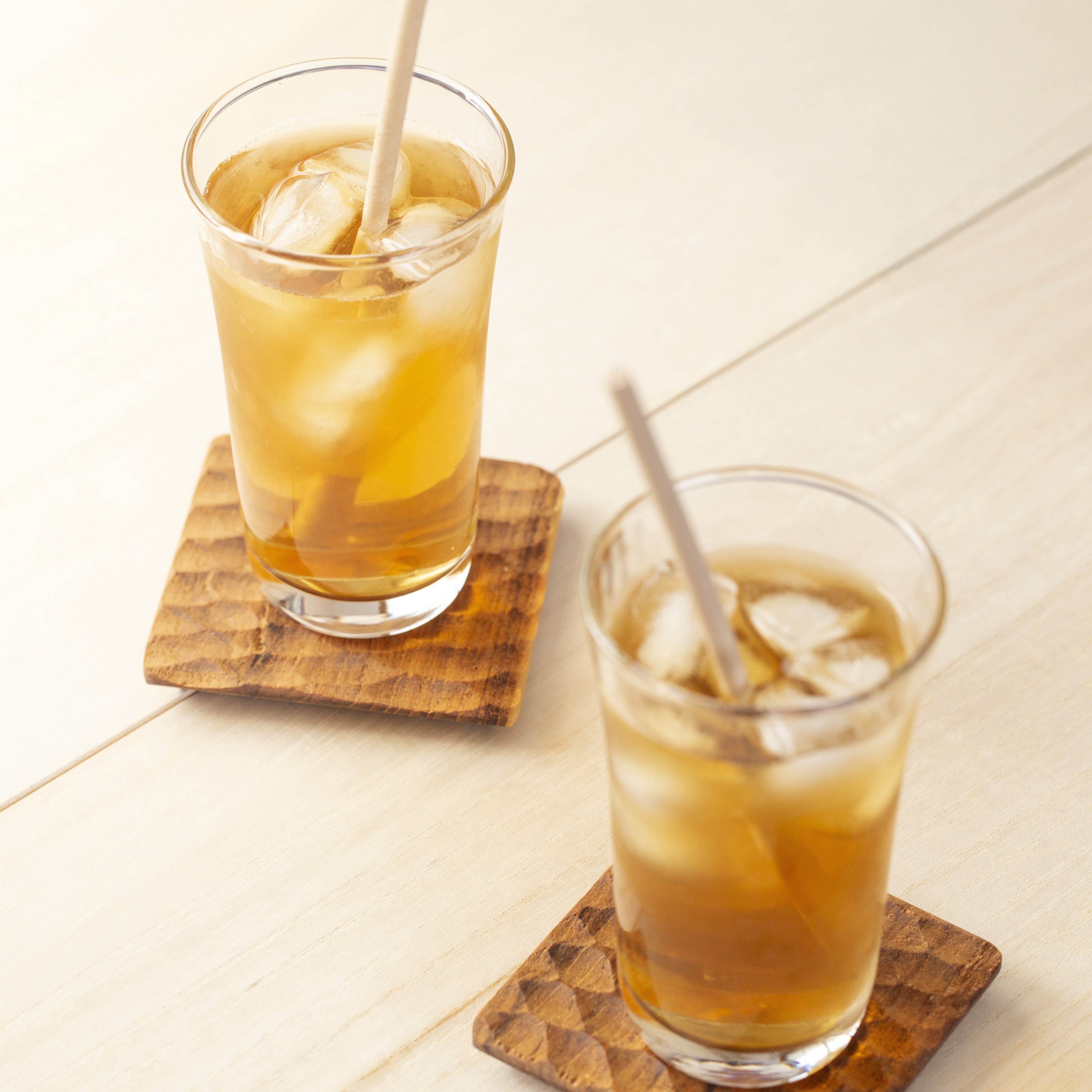 Two glasses of iced tea with straws on wooden coasters.