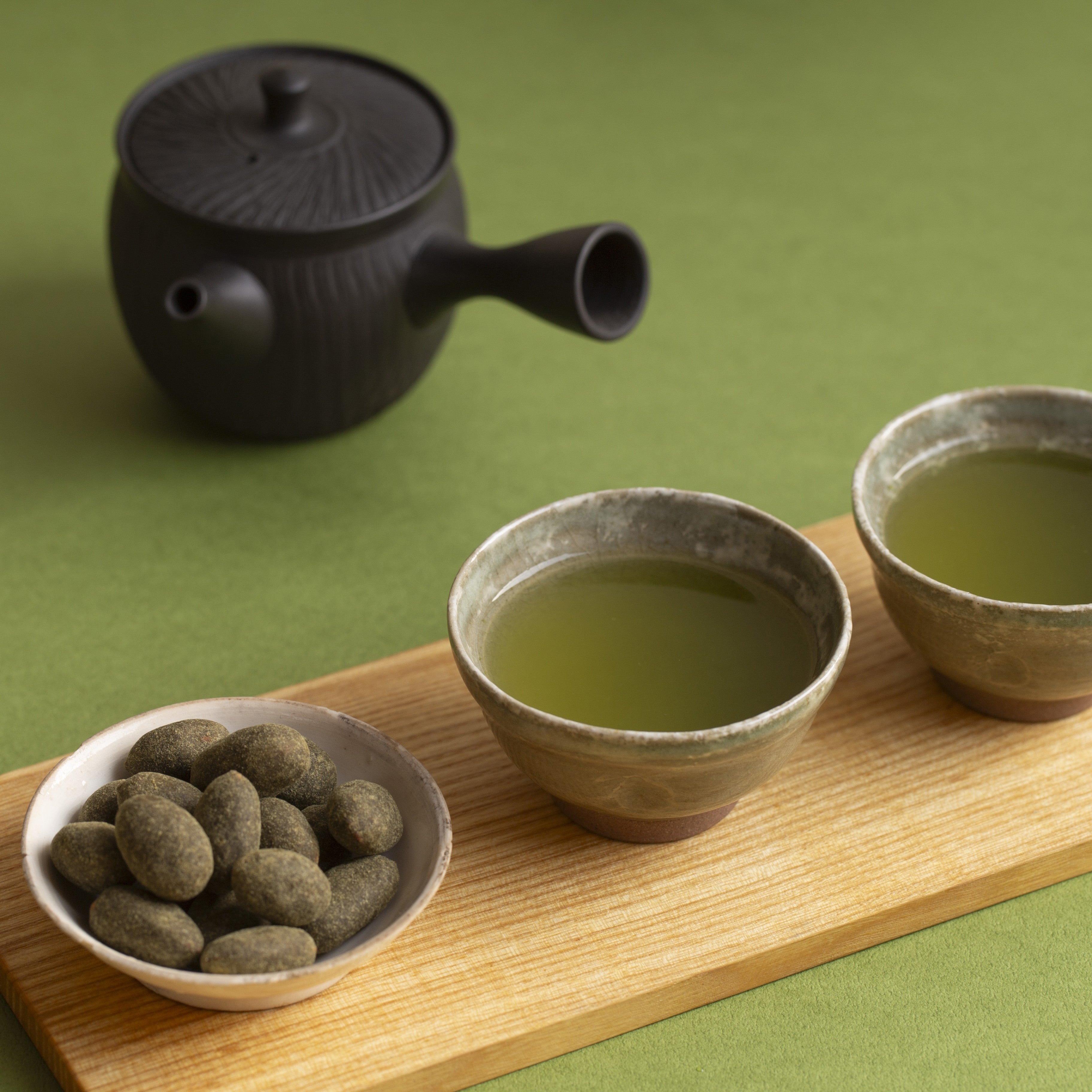 Two bowls of green tea with a black teapot on a wooden tray against a green background
