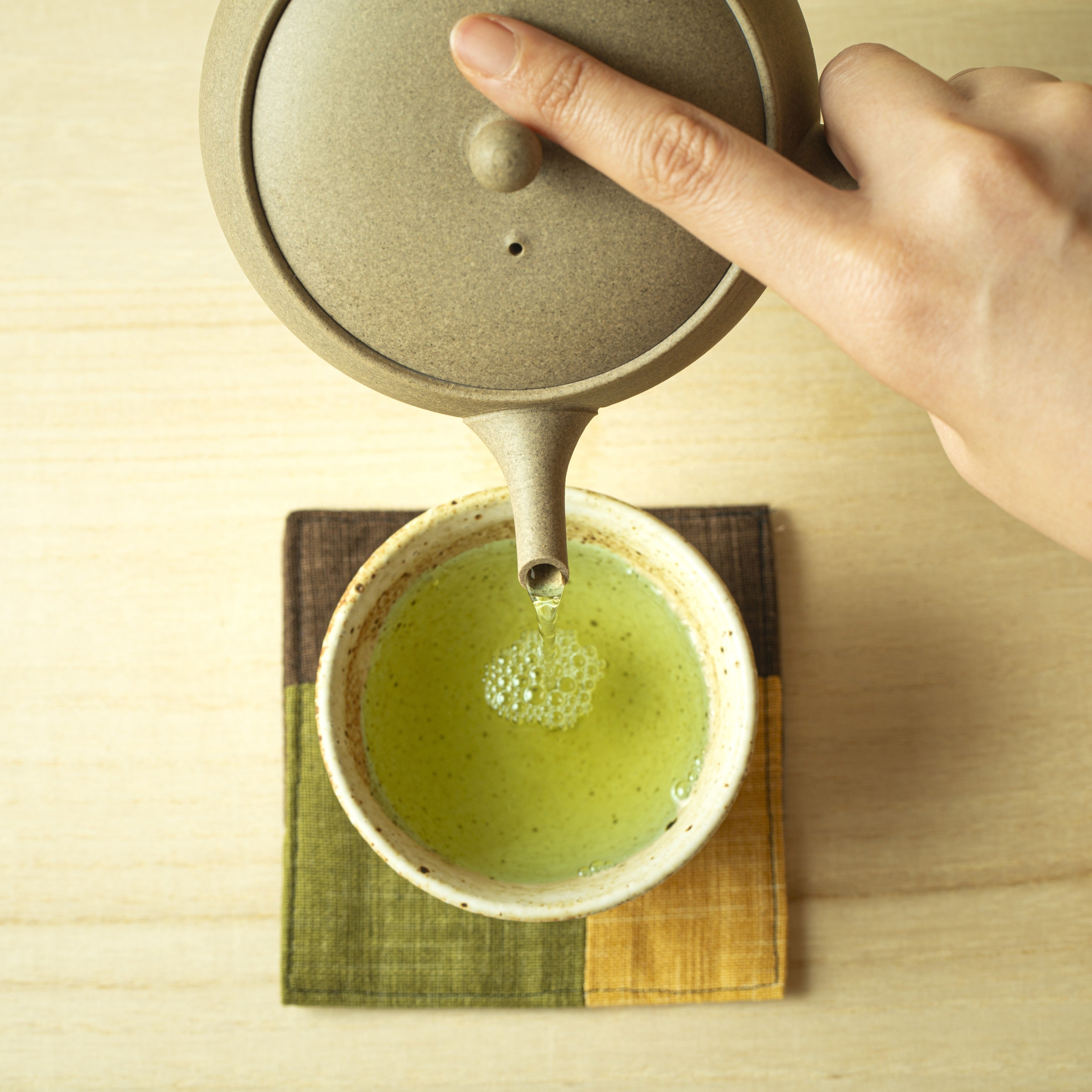 Hand pouring green tea from a teapot into a cup on a wooden surface.