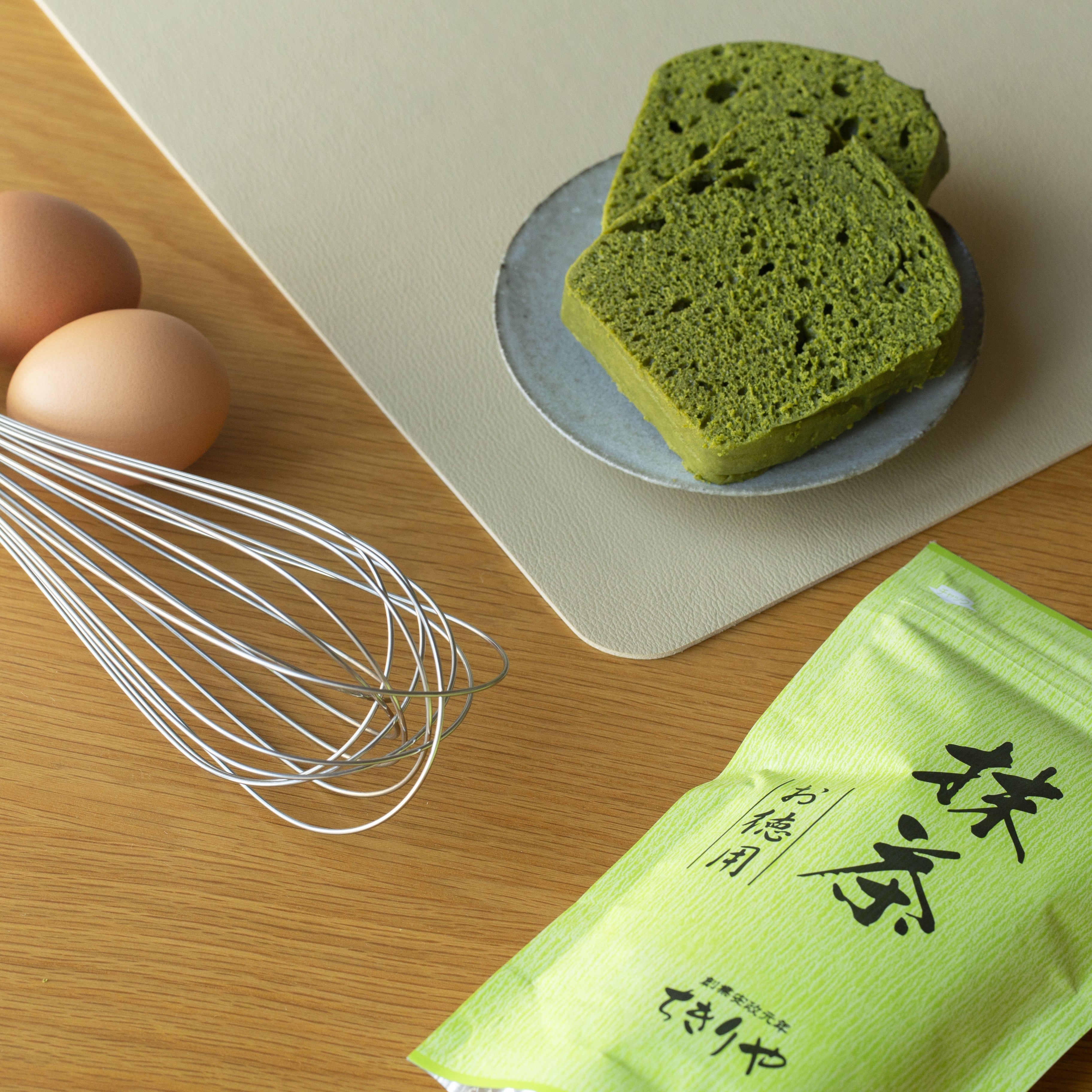 Green matcha cake on a plate with eggs, a whisk and matcha packaging on a wooden surface