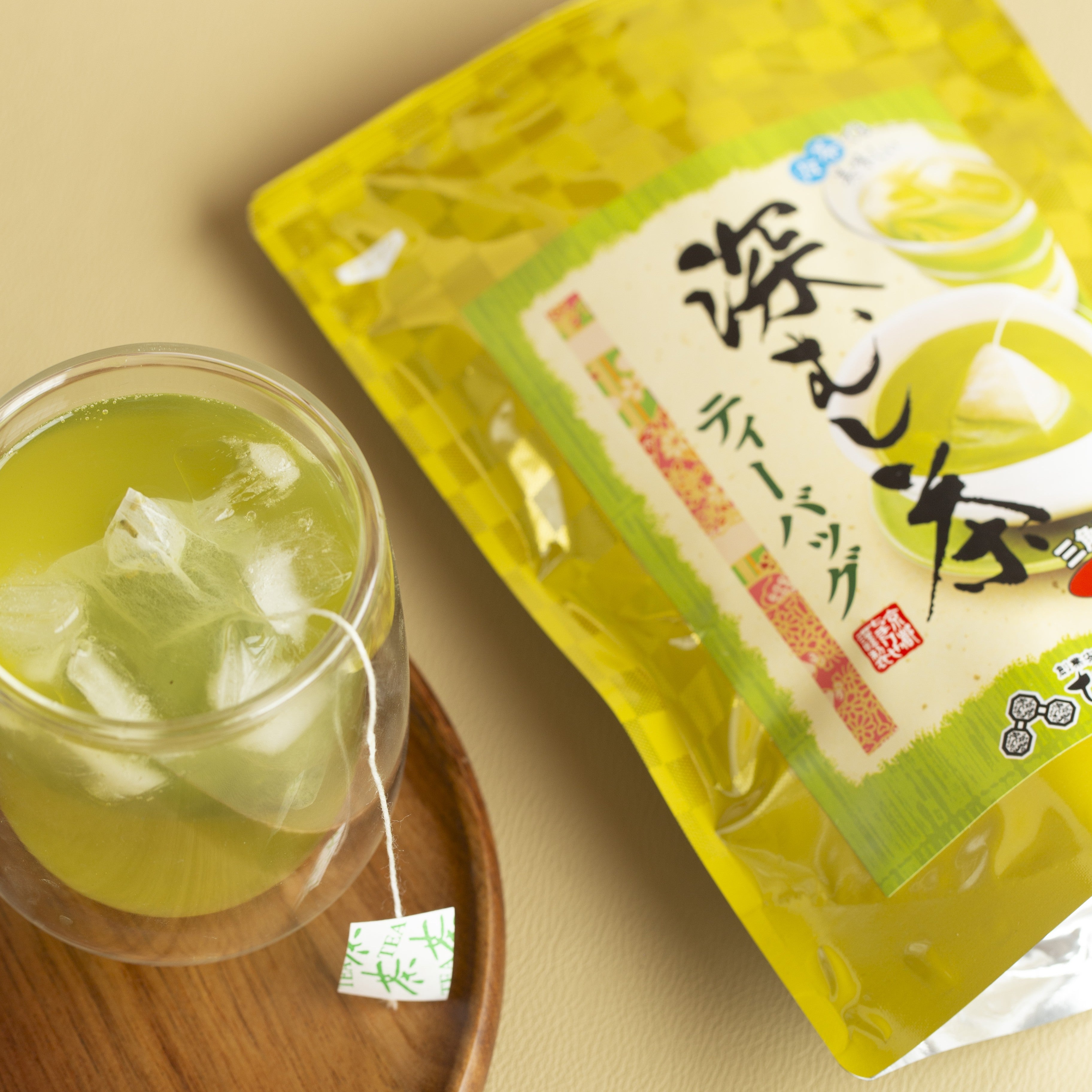 Green tea bag in a glass of green tea next to a yellow package with Japanese text on a beige background