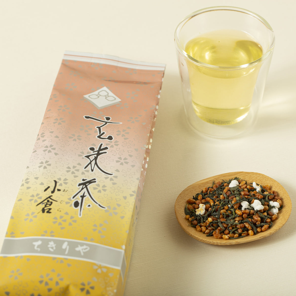 Genmaicha packaging, glass of tea, and wooden dish with tea leaves on a light background
