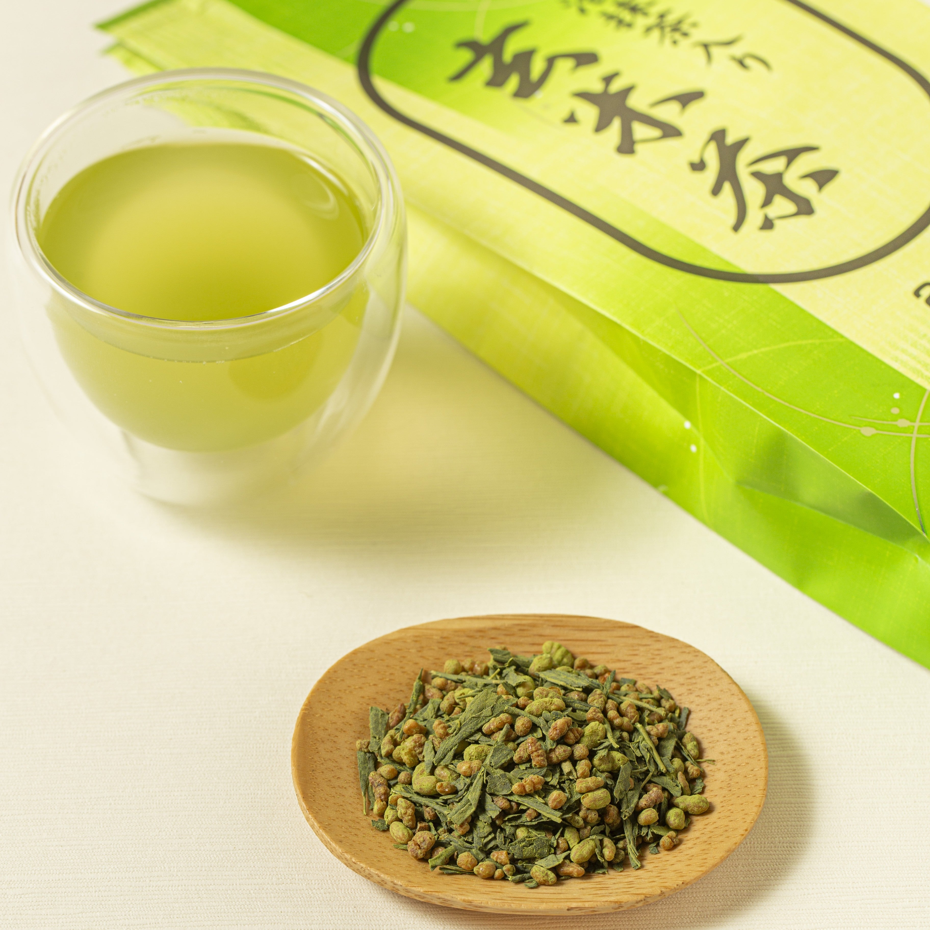 Green tea packaging, glass of green tea, and wooden plate with tea leaves on a light background