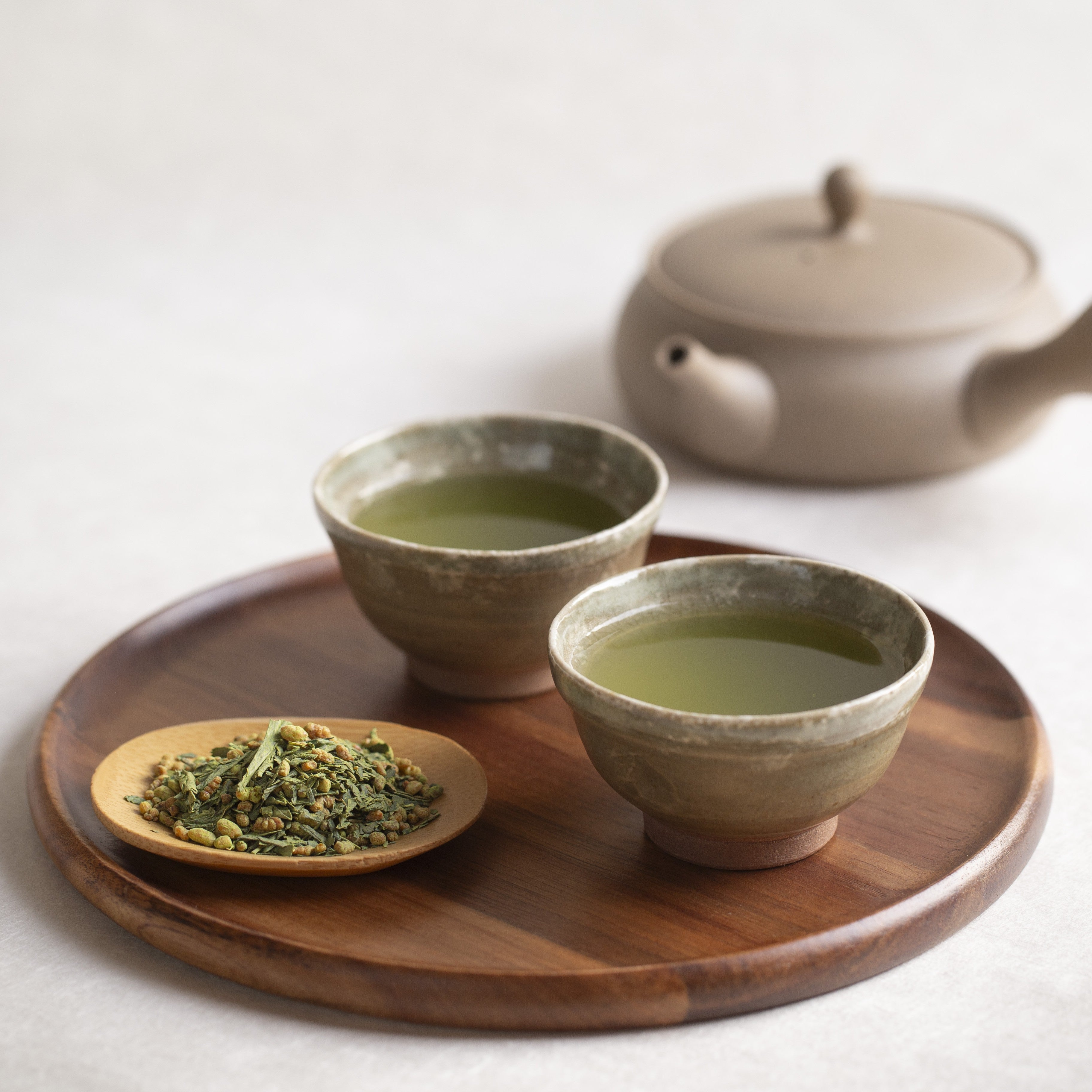Two ceramic bowls with genmaicha matcha green tea on a wooden tray, accompanied by a teapot and a small dish of tea leaves.
