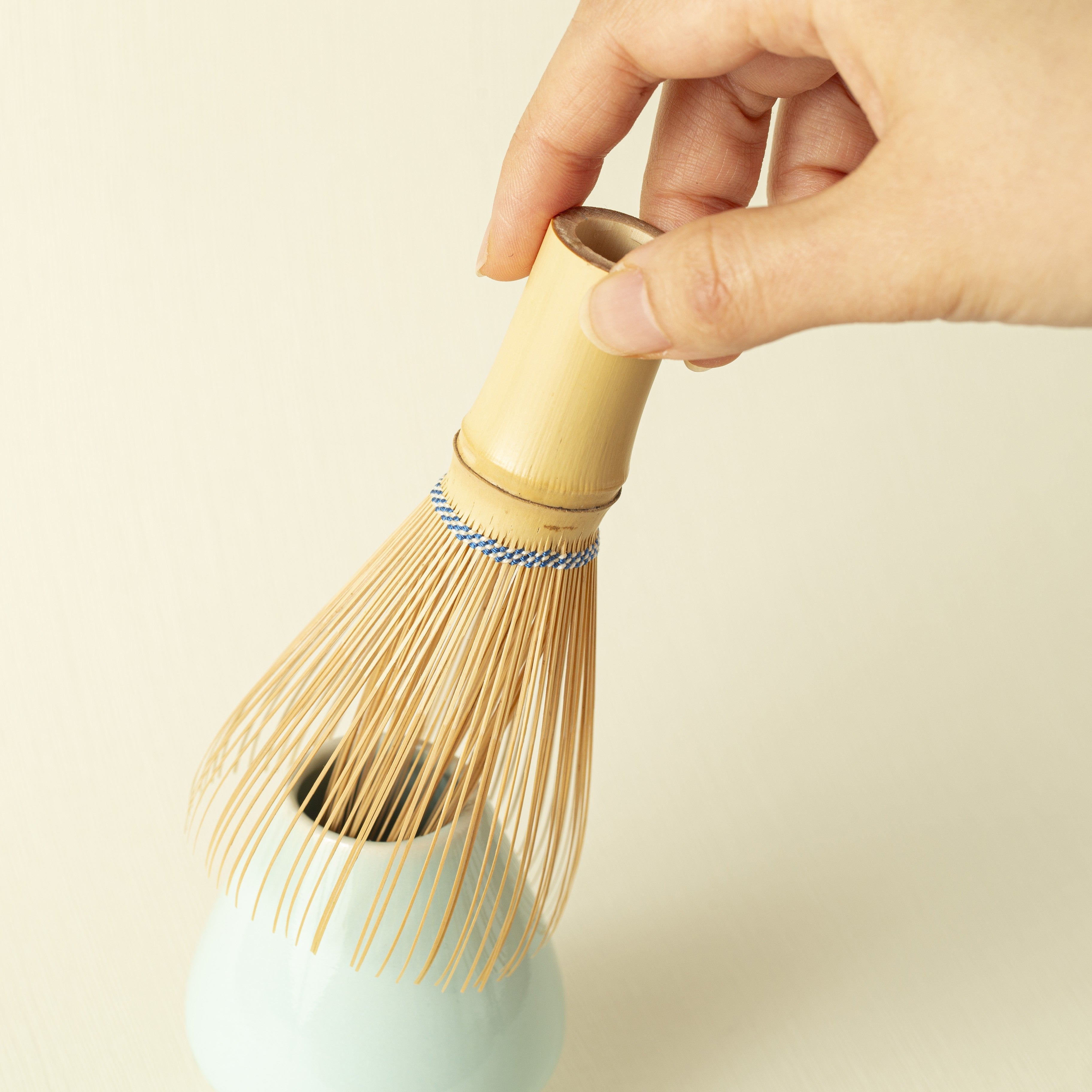 Hand holding a traditional Japanese tea whisk on a blue Japanese whisk stand with a light background