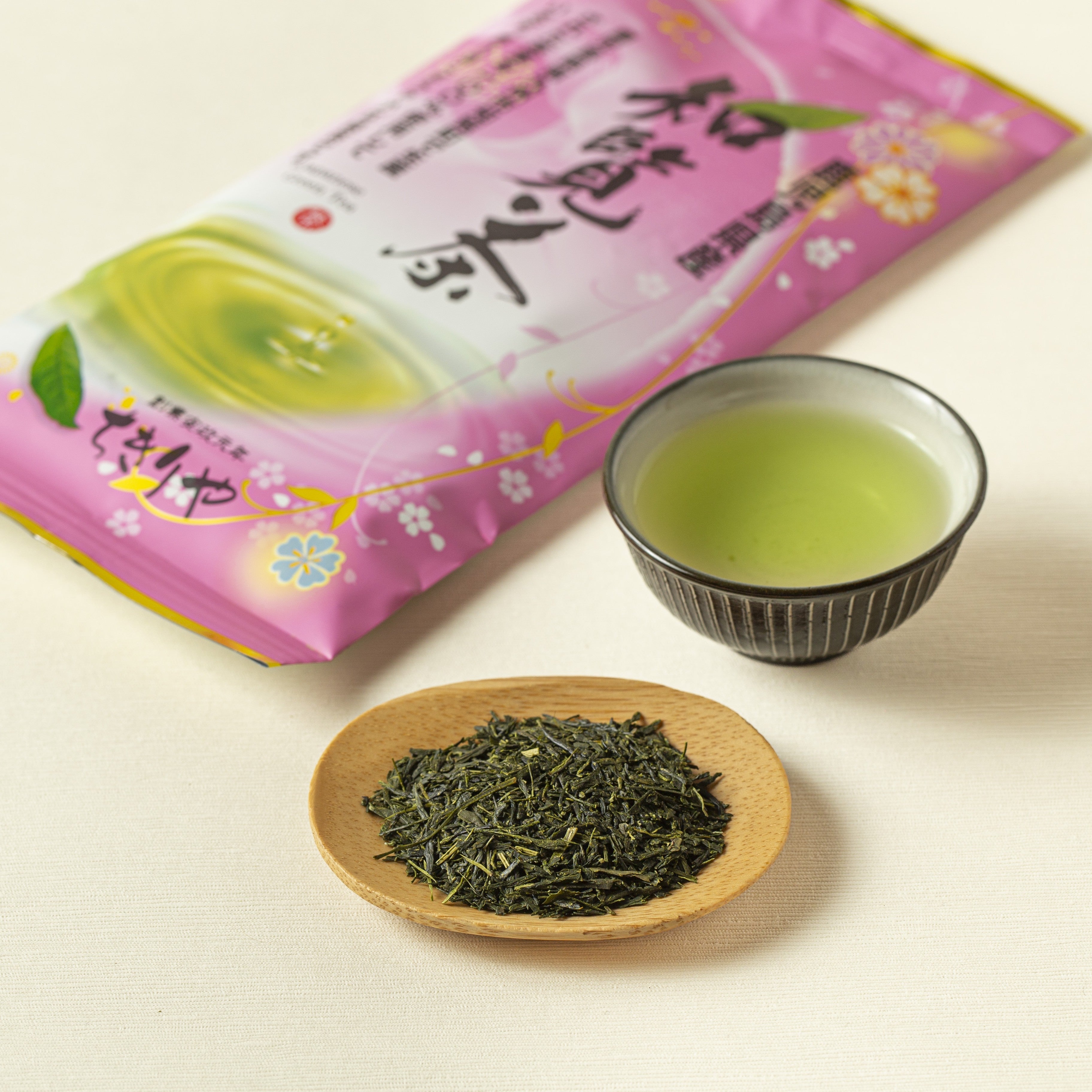 Green tea leaves on a wooden plate with a cup of green tea and packaging in the background.