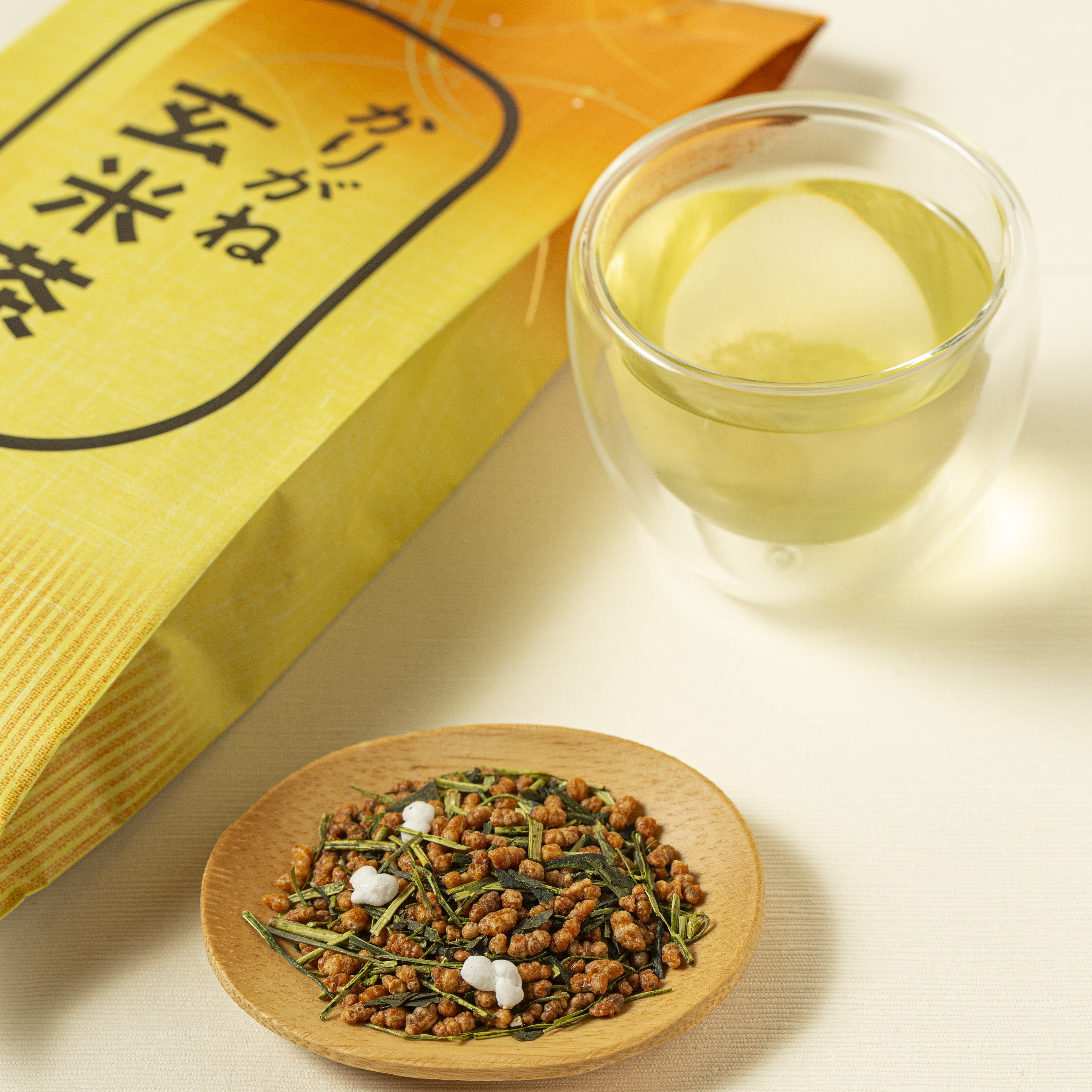 Tea leaves on a wooden plate with a glass of tea and yellow packaging in the background.