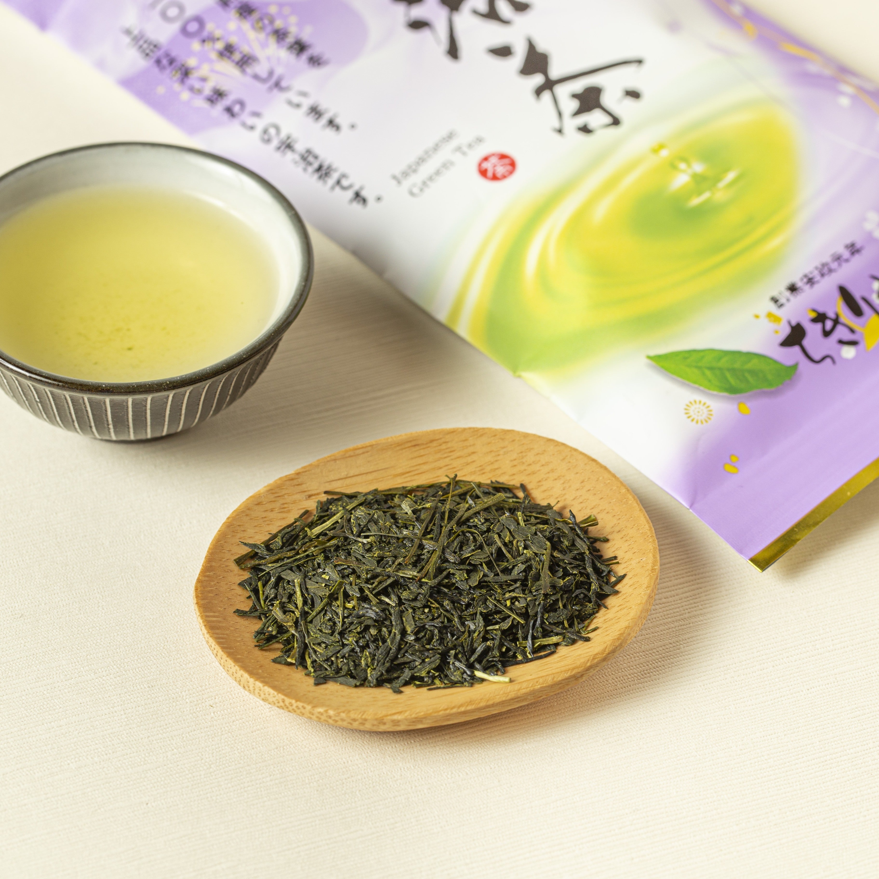 Tea leaves on a wooden plate with a bowl of tea and packaging in the background.