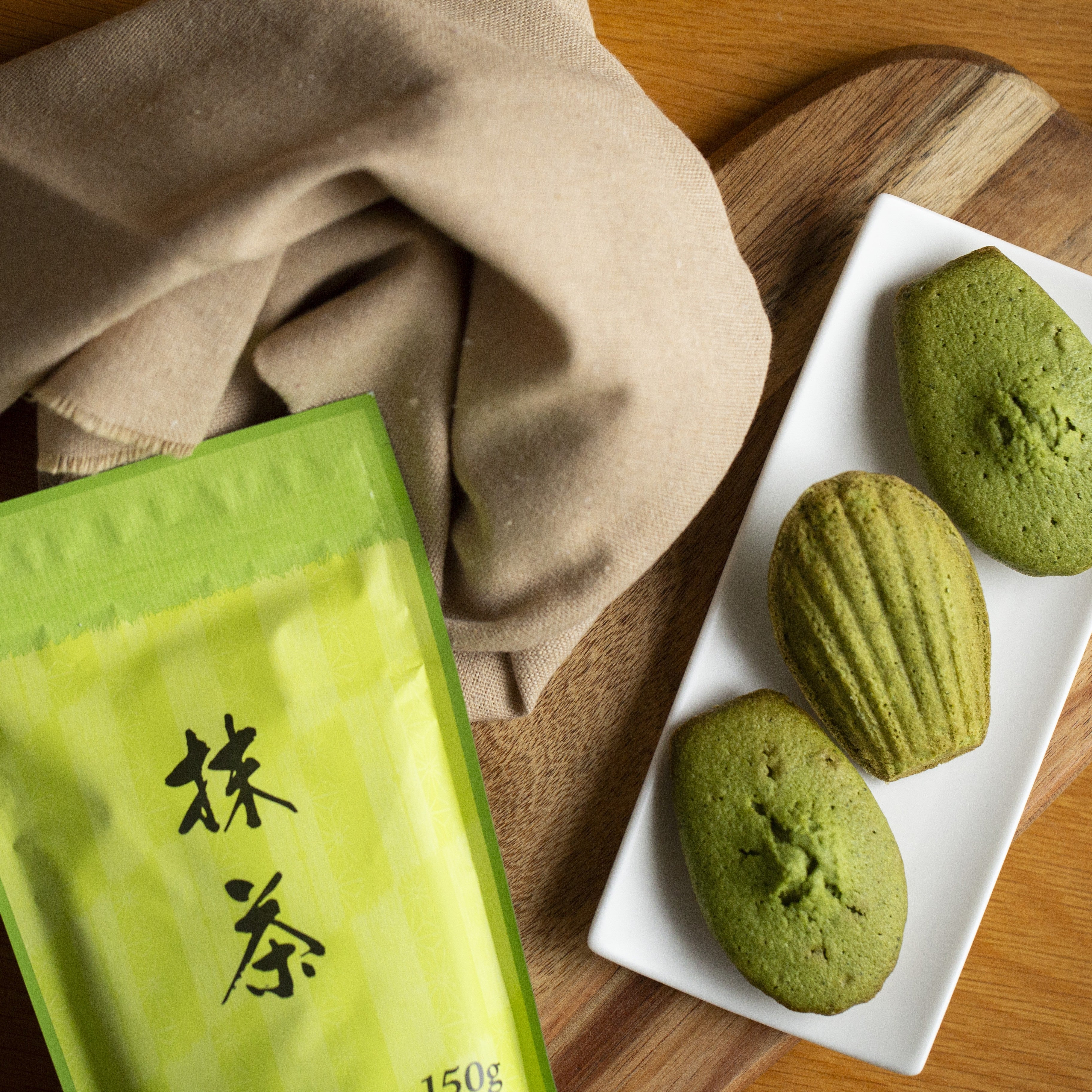 Green tea package and matcha madeleineson a wooden board with a beige cloth