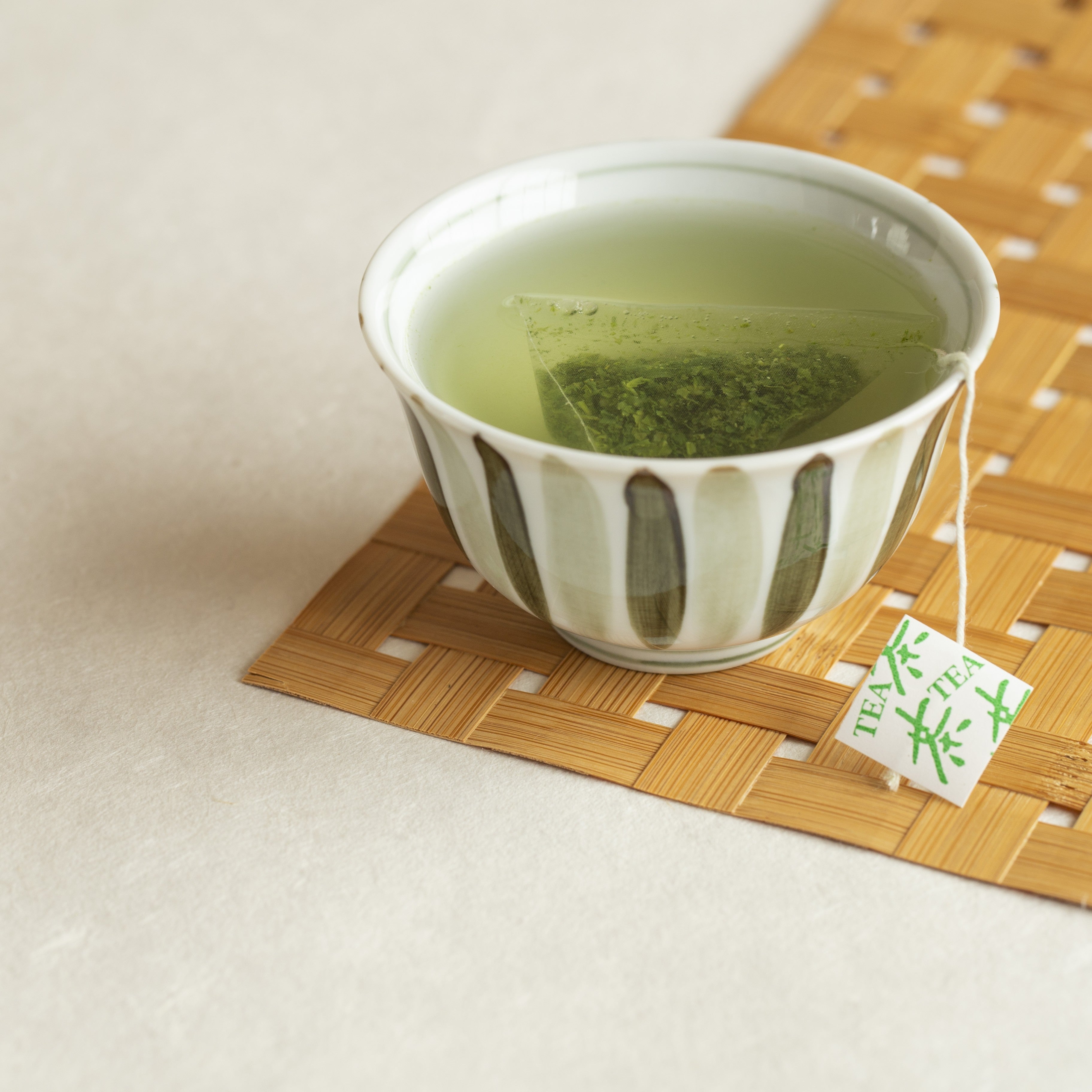 Green tea in a striped cup on a bamboo mat with a tea bag tag.