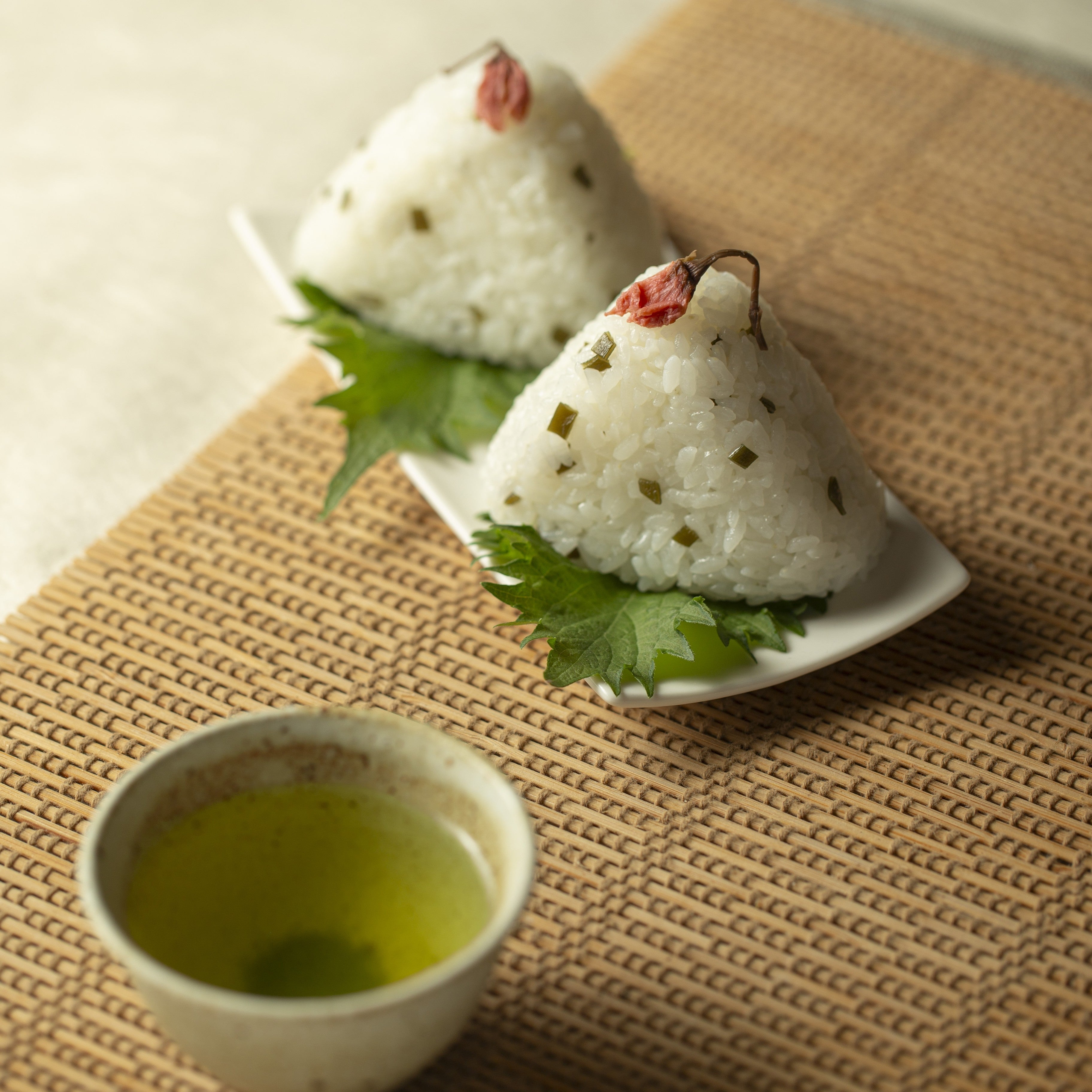 Two white rice balls with green leaves on a white plate, accompanied by a small bowl of green tea on a woven mat.